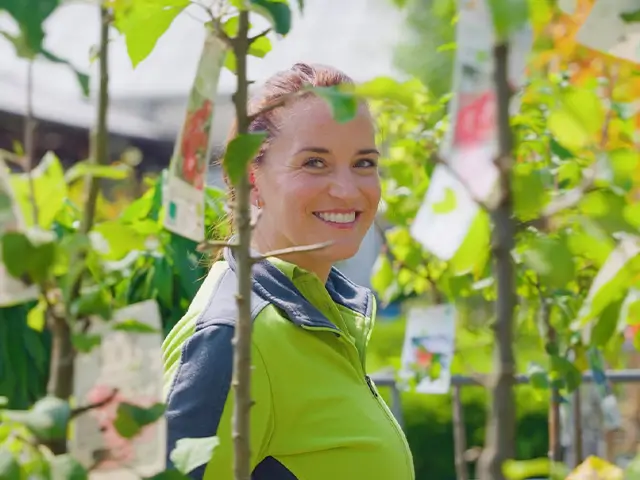 Eine Frau lächelt zwischen jungen Bäumen im Gartencenter Augsburg.